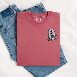 May include: A maroon t-shirt with a white ghost holding a red can. The t-shirt has a small tag that says "COMFORT COLORS" on it.