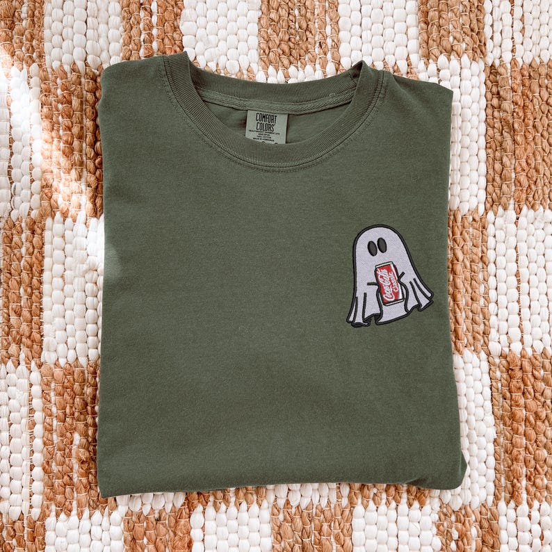 Op de afbeelding: Een olijfgroen t-shirt met ronde hals en een geborduurde patch op de linkerborst. De patch toont een lichtgrijze geest met zwarte ogen die een klein rood Coca-Cola-blikje vasthoudt. Het shirt heeft een Comfort Colors label in de nek.