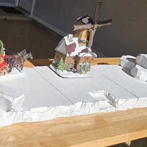 May include: A Christmas village display featuring a red wagon with a sign that reads "Christmas Tree", pulled by two brown horses. A windmill and a house are also part of the display, along with a pile of white rocks.