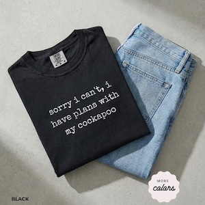 Peut inclure: Un t-shirt noir à manches courtes avec le texte imprimé en blanc 'sorry i can't, i have plans with my cockapoo' associé à un jean bleu clair. Le t-shirt présente une encolure ronde et une étiquette de marque visible au col intérieur.