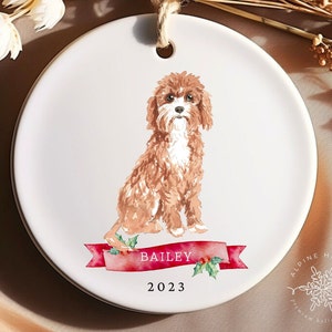 Op de afbeelding: Een ronde, witte keramische kerstversiering met een aquarel illustratie van een bruine en witte hond. De naam van de hond, "Bailey", staat op een rode banner met hulst, en het jaar "2023" staat eronder. Een jute touwtje is bevestigd.