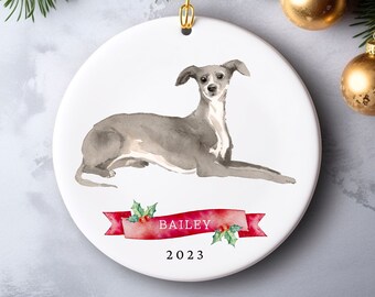 May include: White ceramic Christmas ornament featuring a watercolor illustration of a greyhound dog. The dog is lying down. A red banner with the name "BAILEY" and the year "2023" is below the dog. The ornament has a gold hanging loop.