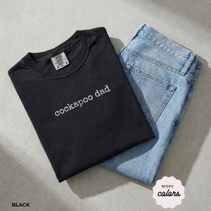 Puede incluir: Una camiseta negra de manga corta con el texto blanco "cockapoo dad" impreso en el pecho, combinada con unos vaqueros de mezclilla azul claro. La camiseta presenta un cuello redondo y una etiqueta de la marca Comfort Colors visible en el interior.