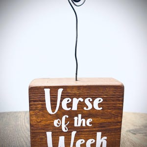 May include: A wooden block with the text "Verse of the Week" in white lettering. A wire spiral extends from the top of the block. The block is brown and sits on a wooden surface.