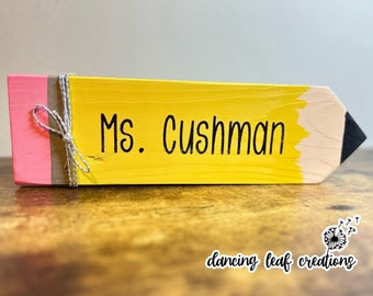 May include: A yellow wooden pencil with a pink eraser and a gray twine bow. The pencil is painted with the text "Ms. Cushman".