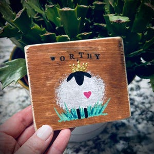 May include: A square wooden plaque with the word "WORTHY" in black letters. It features a hand-painted sheep wearing a gold crown and a pink heart. The sheep is on green grass. The plaque is held by a person.