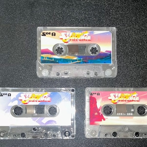 Puede incluir: Tres cintas de cassette transparentes con el logotipo de "Steven Universe" e ilustraciones coloridas. Cada cinta tiene un dise&ntilde;o de fondo diferente, incluyendo rosa, azul y una escena de puesta de sol. La etiqueta dice "Lado A".