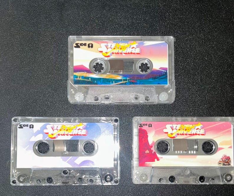 Puede incluir: Tres cintas de cassette transparentes con el logotipo de "Steven Universe" e ilustraciones coloridas. Cada cinta tiene un dise&ntilde;o de fondo diferente, incluyendo rosa, azul y una escena de puesta de sol. La etiqueta dice "Lado A".