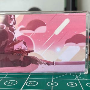 Puede incluir: Una funda transparente para casete con una ilustraci&oacute;n rosa y roja. La obra de arte representa una figura, una casa en un acantilado, nubes y estrellas fugaces contra un cielo de atardecer. La funda est&aacute; sobre una esterilla de corte verde.