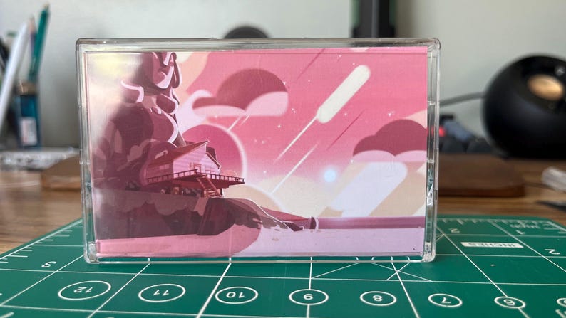 Puede incluir: Una funda transparente para casete con una ilustraci&oacute;n rosa y roja. La obra de arte representa una figura, una casa en un acantilado, nubes y estrellas fugaces contra un cielo de atardecer. La funda est&aacute; sobre una esterilla de corte verde.