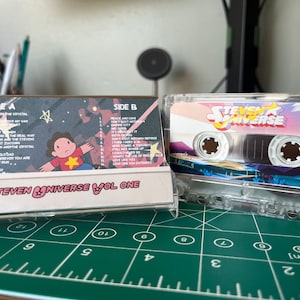 May include: A clear cassette tape labeled "Steven Universe Vol. One" with a colorful design. The cassette case shows the track list and a cartoon character. The tape is on a green cutting mat.
