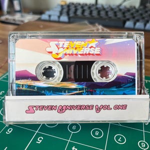 Puede incluir: Una cinta de cassette transparente con el logotipo y la ilustraci&oacute;n de "Steven Universe". La caja del cassette tiene el texto "Steven Universe Vol One" en la parte inferior. La etiqueta de la cinta muestra un paisaje colorido con el t&iacute;tulo del programa.
