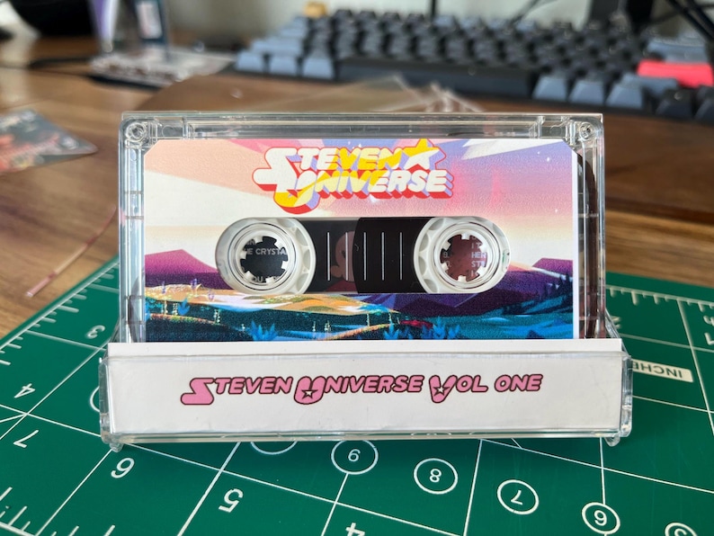 Puede incluir: Una cinta de cassette transparente con el logotipo y la ilustraci&oacute;n de "Steven Universe". La caja del cassette tiene el texto "Steven Universe Vol One" en la parte inferior. La etiqueta de la cinta muestra un paisaje colorido con el t&iacute;tulo del programa.