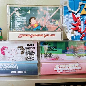 Puede incluir: Una colecci&oacute;n de tres cintas de casete de Steven Universe. Las cintas est&aacute;n etiquetadas como "Volume One", "Volume 2" y "The Movie". Las cintas de casete tienen ilustraciones y texto coloridos, incluidos t&iacute;tulos de canciones e ilustraciones de personajes. Las cintas est&aacute;n apiladas.