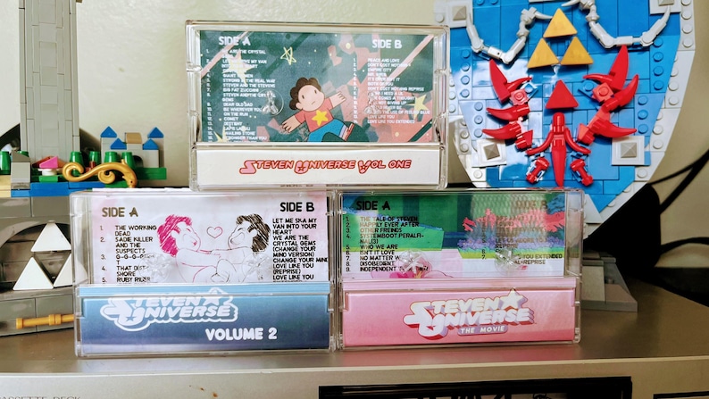 Puede incluir: Una colecci&oacute;n de tres cintas de casete de Steven Universe. Las cintas est&aacute;n etiquetadas como "Volume One", "Volume 2" y "The Movie". Las cintas de casete tienen ilustraciones y texto coloridos, incluidos t&iacute;tulos de canciones e ilustraciones de personajes. Las cintas est&aacute;n apiladas.
