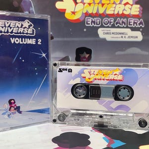 Puede incluir: Una cinta de cassette transparente y su estuche con el logotipo y la ilustraci&oacute;n de "Steven Universe". La cinta est&aacute; etiquetada como "Cara A" y tiene un dise&ntilde;o colorido. Se ve un segundo estuche, con la etiqueta "Volumen 2".