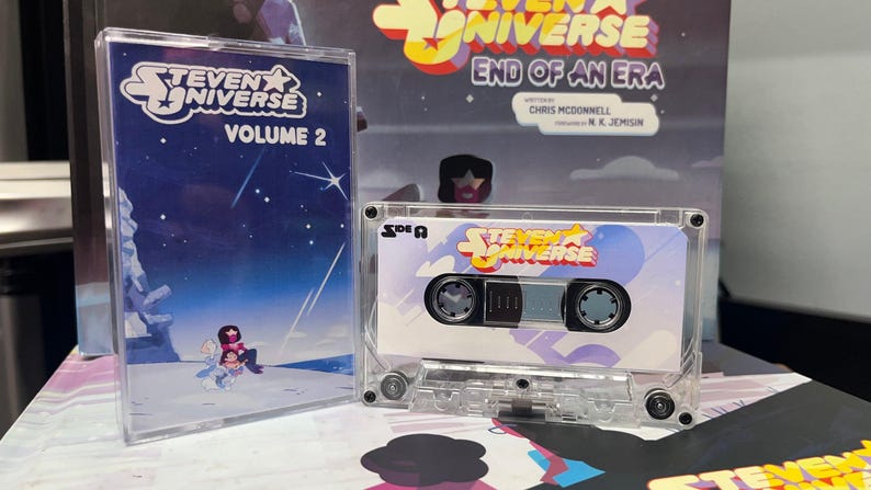 Puede incluir: Una cinta de cassette transparente y su estuche con el logotipo y la ilustraci&oacute;n de "Steven Universe". La cinta est&aacute; etiquetada como "Cara A" y tiene un dise&ntilde;o colorido. Se ve un segundo estuche, con la etiqueta "Volumen 2".