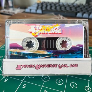 May include: A clear cassette tape case with a Steven Universe design. The cassette tape features the show's logo and a colorful landscape. The bottom of the case reads "Steven Universe Vol One."
