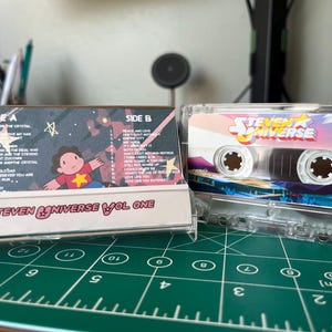 Puede incluir: Una cinta de cassette transparente con ilustraciones de la serie animada "Steven Universe". La caja del cassette muestra el logotipo del programa y un paisaje colorido. La etiqueta del cassette enumera los t&iacute;tulos de las canciones e ilustraciones de personajes.
