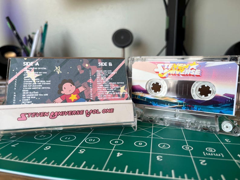 Puede incluir: Una cinta de cassette transparente con ilustraciones de la serie animada "Steven Universe". La caja del cassette muestra el logotipo del programa y un paisaje colorido. La etiqueta del cassette enumera los t&iacute;tulos de las canciones e ilustraciones de personajes.