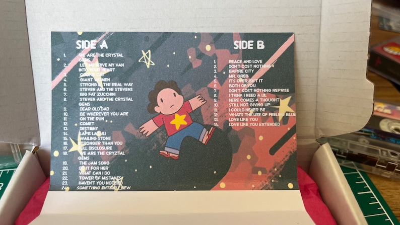 May include: A cassette tape insert featuring a cartoon character and song titles. The design has a space theme with stars and a color palette of red, blue, and purple. The text includes song titles from Side A and Side B.