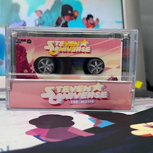 Puede incluir: Un coche de juguete rosa y blanco con el tema de Steven Universe, en una vitrina transparente. El coche tiene ruedas negras y el logotipo de Steven Universe. La caja tambi&eacute;n tiene el texto "Steven Universe The Movie".
