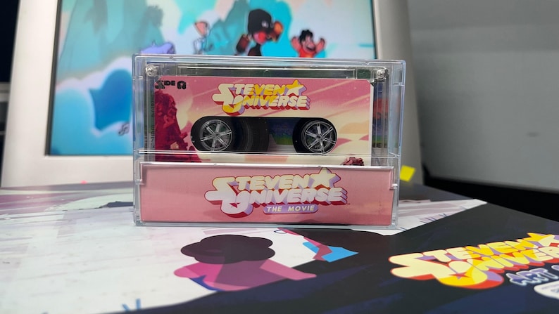 Puede incluir: Un coche de juguete rosa y blanco con el tema de Steven Universe, en una vitrina transparente. El coche tiene ruedas negras y el logotipo de Steven Universe. La caja tambi&eacute;n tiene el texto "Steven Universe The Movie".