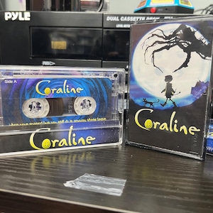 May include: A clear cassette tape and case featuring the movie title "Coraline" in yellow script. The cassette tape has a blue and purple design. The case shows a silhouette of a child and cat in front of a full moon.