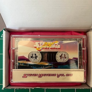 May include: A cassette tape featuring the "Steven Universe" logo and artwork, encased in a clear plastic case. The cassette is labeled "Steven Universe Vol One" and is presented in a white box with a pink interior.
