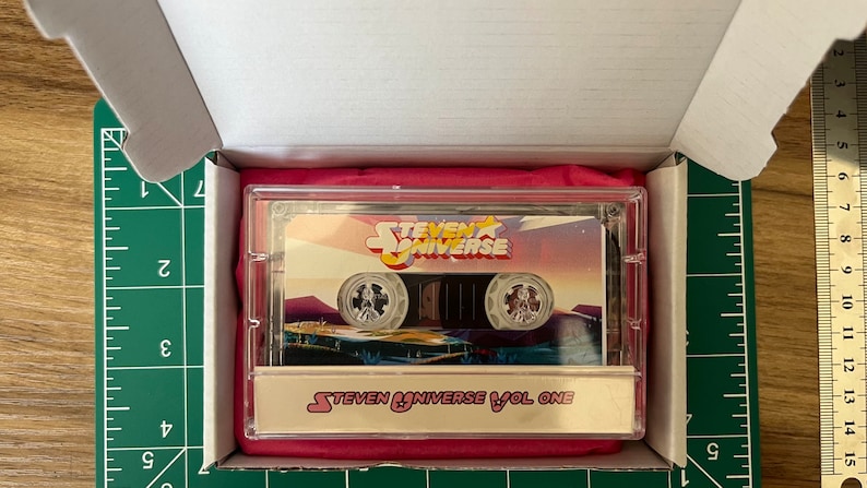 May include: A cassette tape featuring the "Steven Universe" logo and artwork, encased in a clear plastic case. The cassette is labeled "Steven Universe Vol One" and is presented in a white box with a pink interior.