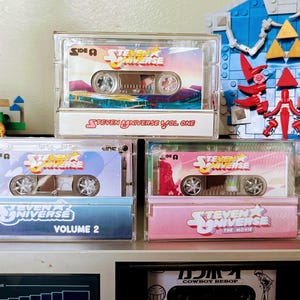 Puede incluir: Tres cintas de cassette de Steven Universe en estuches transparentes est&aacute;n apiladas. La cinta superior dice "Steven Universe Vol. One". La cinta del medio dice "Volume 2". La cinta inferior dice "The Movie". Un escudo de Lego est&aacute; en el fondo.