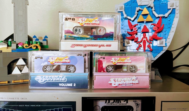 Puede incluir: Tres cintas de cassette de Steven Universe en estuches transparentes est&aacute;n apiladas. La cinta superior dice "Steven Universe Vol. One". La cinta del medio dice "Volume 2". La cinta inferior dice "The Movie". Un escudo de Lego est&aacute; en el fondo.