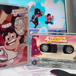 Puede incluir: Una cinta de cassette transparente y su estuche con personajes de la serie animada "Steven Universe". La cinta de cassette tiene una etiqueta rosa y blanca con el logotipo del programa. El estuche muestra una imagen colorida de los personajes.