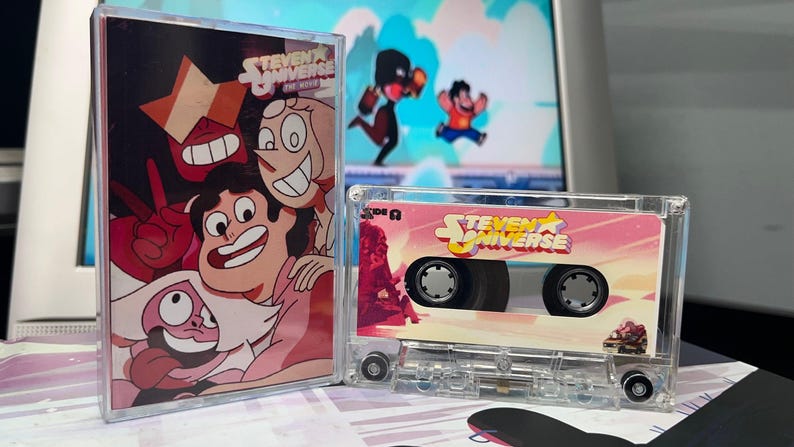 Puede incluir: Una cinta de cassette transparente y su estuche con personajes de la serie animada "Steven Universe". La cinta de cassette tiene una etiqueta rosa y blanca con el logotipo del programa. El estuche muestra una imagen colorida de los personajes.