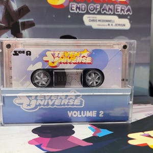 Puede incluir: Una caja de pl&aacute;stico transparente que contiene una cinta de cassette de juguete con el logotipo de "Steven Universe" y el texto "Volume 2". El cassette tiene ruedas plateadas y un centro negro. El fondo incluye una caja con el texto "End of an Era".
