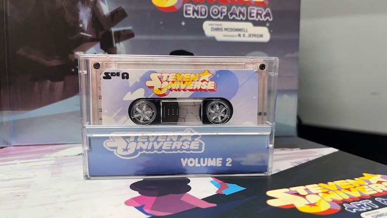 Puede incluir: Una caja de pl&aacute;stico transparente que contiene una cinta de cassette de juguete con el logotipo de "Steven Universe" y el texto "Volume 2". El cassette tiene ruedas plateadas y un centro negro. El fondo incluye una caja con el texto "End of an Era".