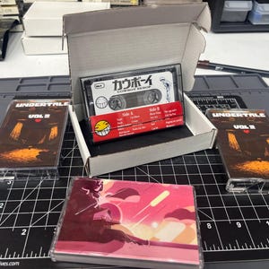 Puede incluir: Tres cintas de casete y una caja de cartón blanca abierta. Dos cintas presentan la ilustración de Undertale Vol 2. Una cinta dentro de la caja muestra el texto Cowboy Bebop y un icono de cara sonriente amarilla. Una cuarta cinta tiene una ilustración.