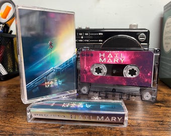 May include: A Project Hail Mary cassette tape with a clear plastic case. The cover art features a space scene with an astronaut and the text Project Hail Mary. The cassette label is pink and purple with the text Side B and Project Hail Mary.