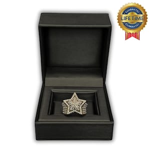 May include: A star-shaped ring with a diamond-like design, presented in a black box. The ring features a large star design with multiple rows of stones. The box is open, revealing the ring. A gold seal with the words "Lifetime Guarantee" is visible.