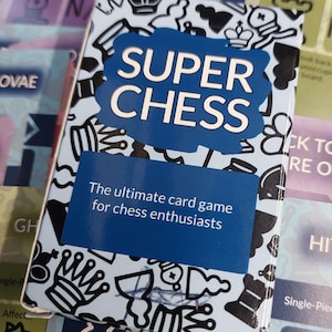 Op de afbeelding: Een kaartspeldoos met de titel "SUPER CHESS" met een blauw en wit ontwerp. De doos bevat illustraties van schaakstukken en de tekst "The ultimate card game for chess enthusiasts." Andere speelkaarten zijn zichtbaar op de achtergrond.