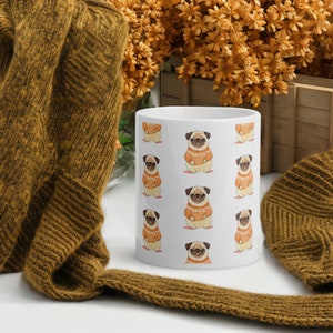 Könnte beinhalten: Weiße Keramiktasse mit einem sich wiederholenden Muster von Mops, die orangefarbene Pullover tragen. Die Möpse blicken nach vorne und haben die Zunge herausgestreckt.