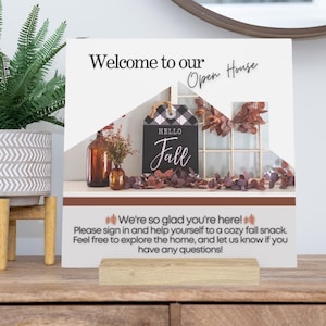 Puede incluir: Un cartel de bienvenida para una casa abierta con temática otoñal. El cartel es blanco con un patrón de cuadros blanco y negro y las palabras "Bienvenido a nuestra casa abierta" en negro. Hay un pequeño cartel que dice "Hola otoño" con una corona y hojas de otoño. El cartel está sobre una mesa de madera con una planta en maceta.