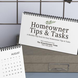 May include: A white flip calendar with the text "Homeowner Tips & Tasks" and the month of August visible. The calendar is on a grey surface. The text "A Monthly Flip Deck for Easy Homeowner Tips & Tasks" is also visible.
