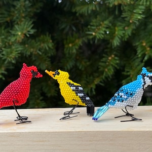May include: Three beaded bird ornaments: a red cardinal, a yellow bird with black detailing, and a blue jay with white and black features. Each bird has a wire base. Green foliage forms the background.