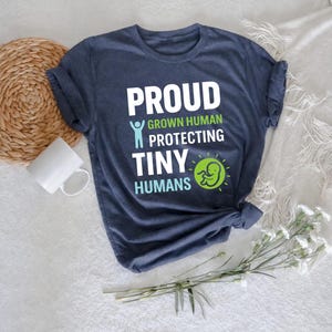 May include: A navy blue t-shirt with the words "PROUD GROWN HUMAN PROTECTING TINY HUMANS" in white and green text. The shirt features a graphic of a person and a baby inside a sun. The shirt is folded and tied at the bottom.