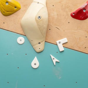 Könnte beinhalten: Eine Sammlung von Klettergriffen, die an einer zweifarbigen Wand montiert sind. Das Set enthält einen großen cremefarbenen Griff, einen gelben, einen roten und mehrere kleinere weiße geometrische Formen, darunter ein kreisförmiges, L-förmiges, tropfenförmiges und pfeilförmiges Teil.