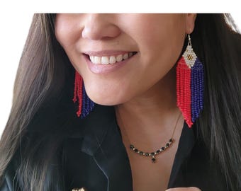 May include: A woman wearing a black shirt and a necklace with black gemstones. She is wearing long, beaded earrings with red, white, and blue colors.