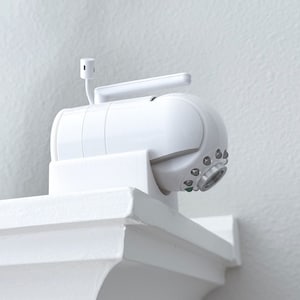 May include: A white baby monitor with a round lens and multiple small black dots around the lens. The monitor is mounted on a white shelf.