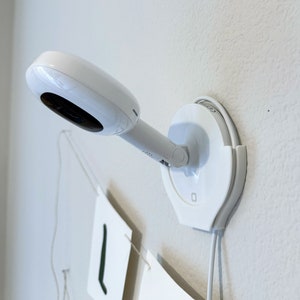 Puede incluir: Un monitor de bebé blanco con una base redonda y una lente de cámara giratoria. El monitor está montado en una pared blanca.