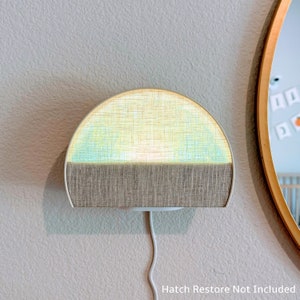 Puede incluir: Una lámpara de pared con forma de semicírculo con una pantalla de tela beige. La mitad superior de la pantalla es de color azul claro. La lámpara está montada en una pared blanca y tiene un cable blanco.